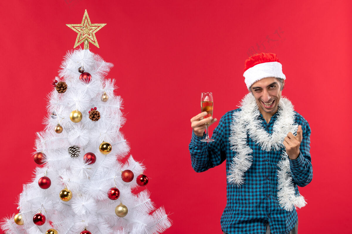 疯狂圣诞心情快乐疯狂情绪好奇的年轻人戴着圣诞老人帽 穿着蓝色条纹衬衫 在圣诞树旁举起一杯葡萄酒年圣诞老人帽子