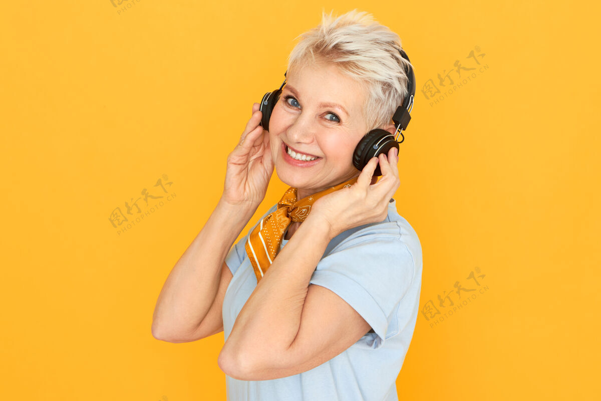 现代时尚的金发退休妇女享受着音乐 戴着无线耳机听收音机摆出一副黄色的姿势耳机欢快穿