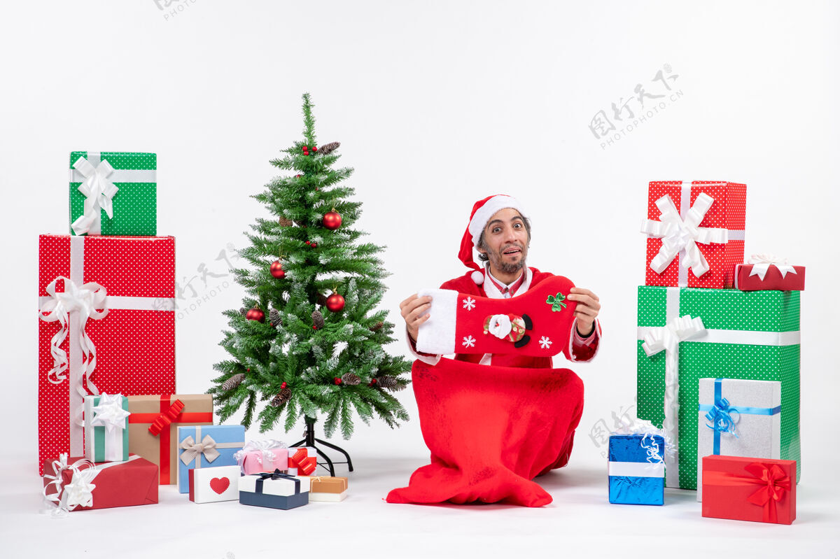 袜子新年气氛有趣积极的圣诞老人坐在地上 拿着圣诞袜子附近的礼物和装饰圣诞树上的白色背景拿着圣诞老人礼物