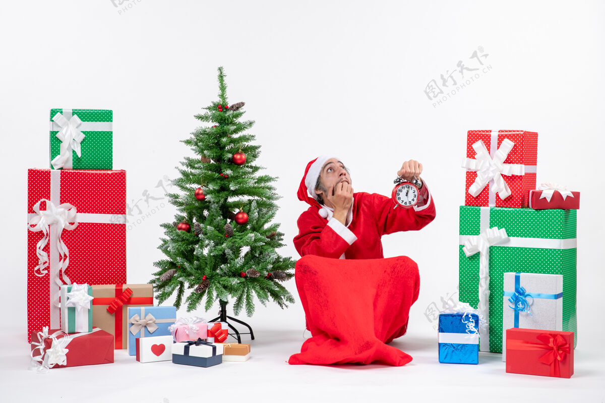 圣诞惊恐的圣诞老人坐在地上沉思着 在礼物和装饰圣诞树旁的白色背景上展示着时钟圣诞老人坐盒子