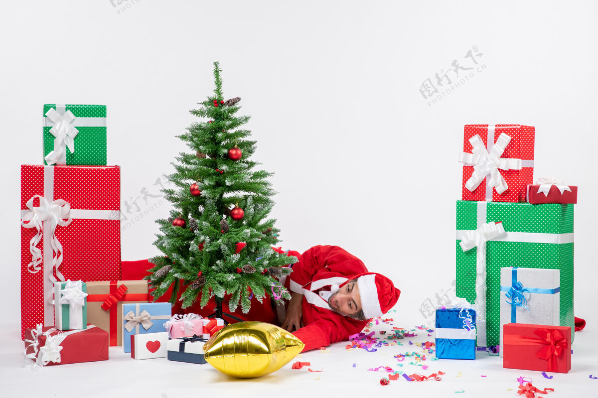 礼物圣诞心情与年轻疲惫的圣诞老人躺在圣诞树后 在白色背景上不同颜色的礼物附近圣诞老人礼物疲倦
