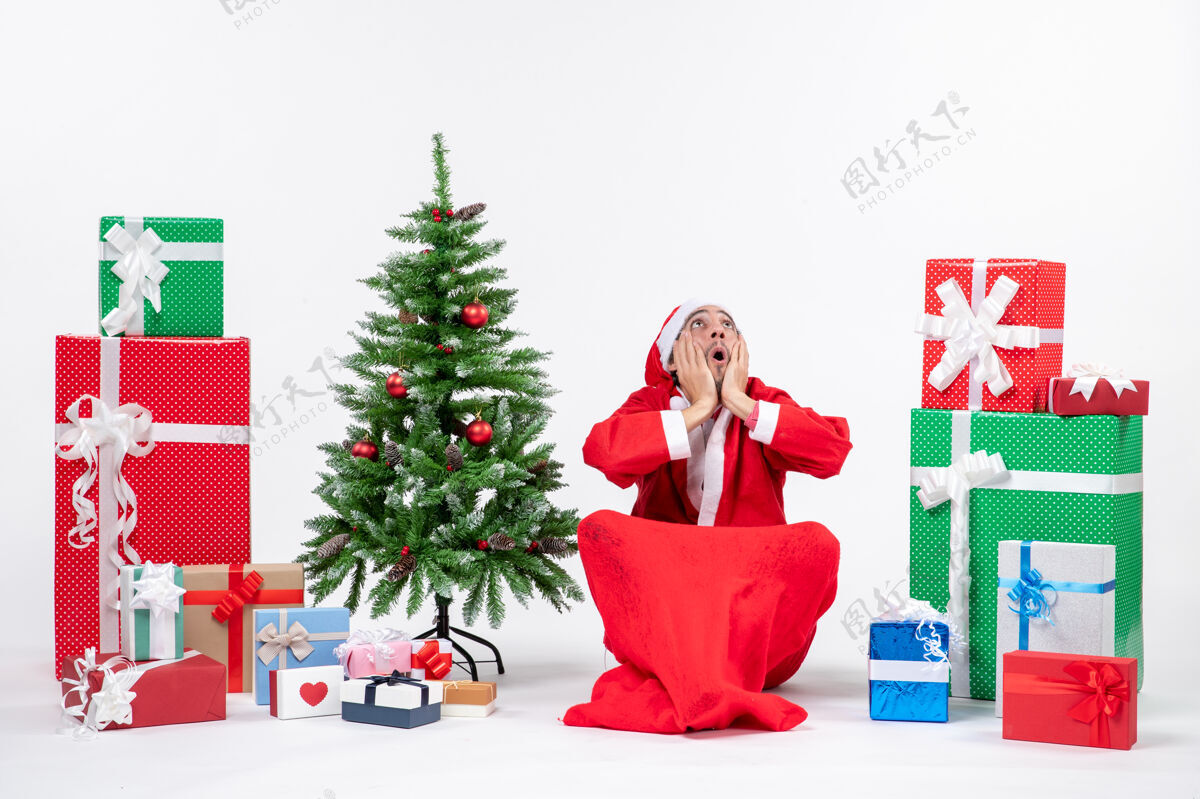 坐着紧张情绪化的年轻人打扮成圣诞老人 拿着礼物和装饰好的圣诞树坐在地上 在白色背景上俯视着地紧张年轻人