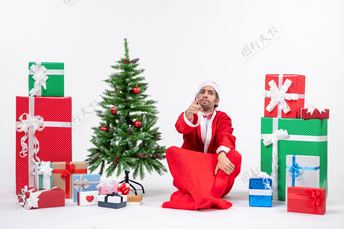 卖家关心的年轻人打扮成圣诞老人与礼物和装饰圣诞树坐在地上的白色背景购物购物者坐着