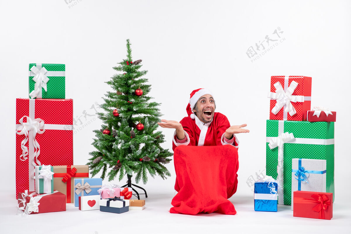 兴奋情绪激动的年轻人打扮成圣诞老人与礼物和装饰圣诞树在白色的背景下反应愉快打扮快乐装饰
