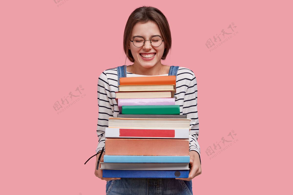 青少年快乐少女的照片拿着一堆教科书 兴高采烈 穿着牛仔工作服 在粉色背景下摆姿势学习学生学习