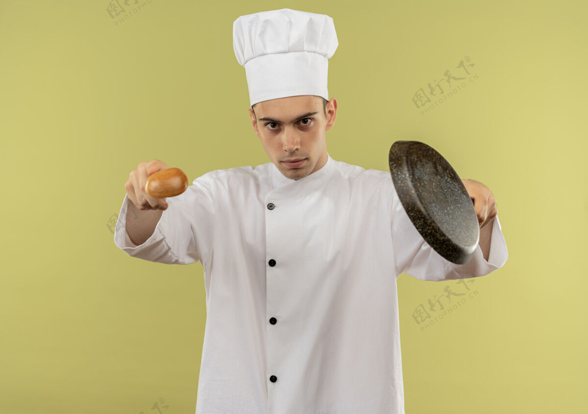 拿着年轻的男厨师穿着厨师制服 在隔离的绿色墙壁上拿着勺子和煎锅制服厨师厨师