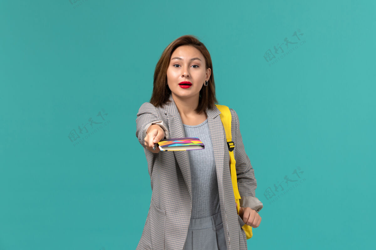 学生身穿灰色夹克 背着黄色背包 拿着文案的女学生在蓝色墙上的正面照片教训夹克人