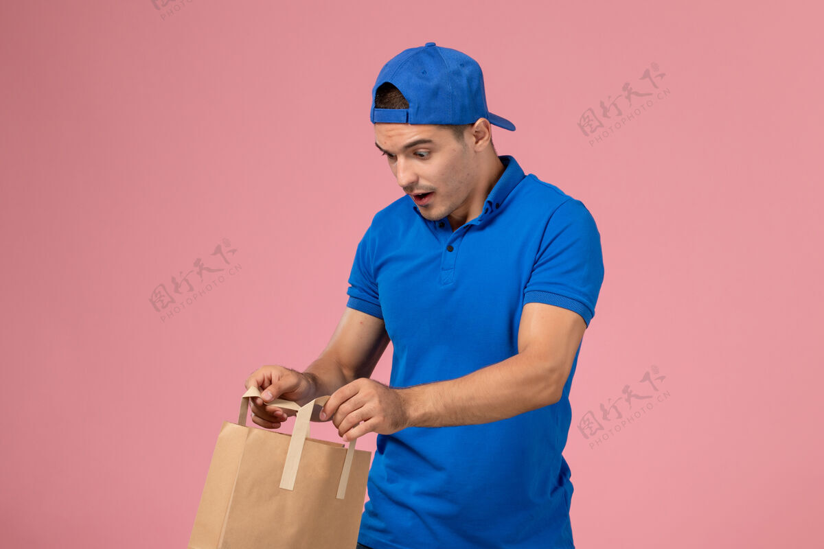 手正面图：身穿蓝色制服和斗篷的年轻男性快递员 手上拿着快递纸包裹 放在粉红色的墙上包裹工作粉色