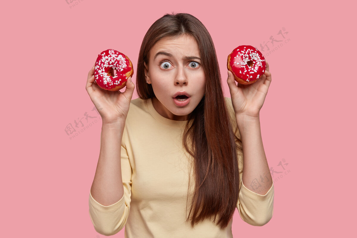 独自尴尬美丽的黑发女人拿着甜甜圈 有惊恐的表情 感到困惑 站在粉色的背景上吃糕点满意