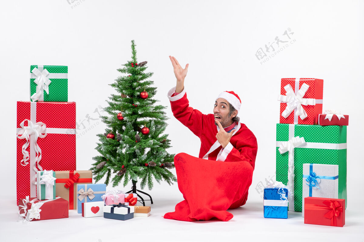 兴奋有趣兴奋的年轻人打扮成圣诞老人与礼物和装饰圣诞树坐在地上指着上面的白色背景成人圣诞老人地上