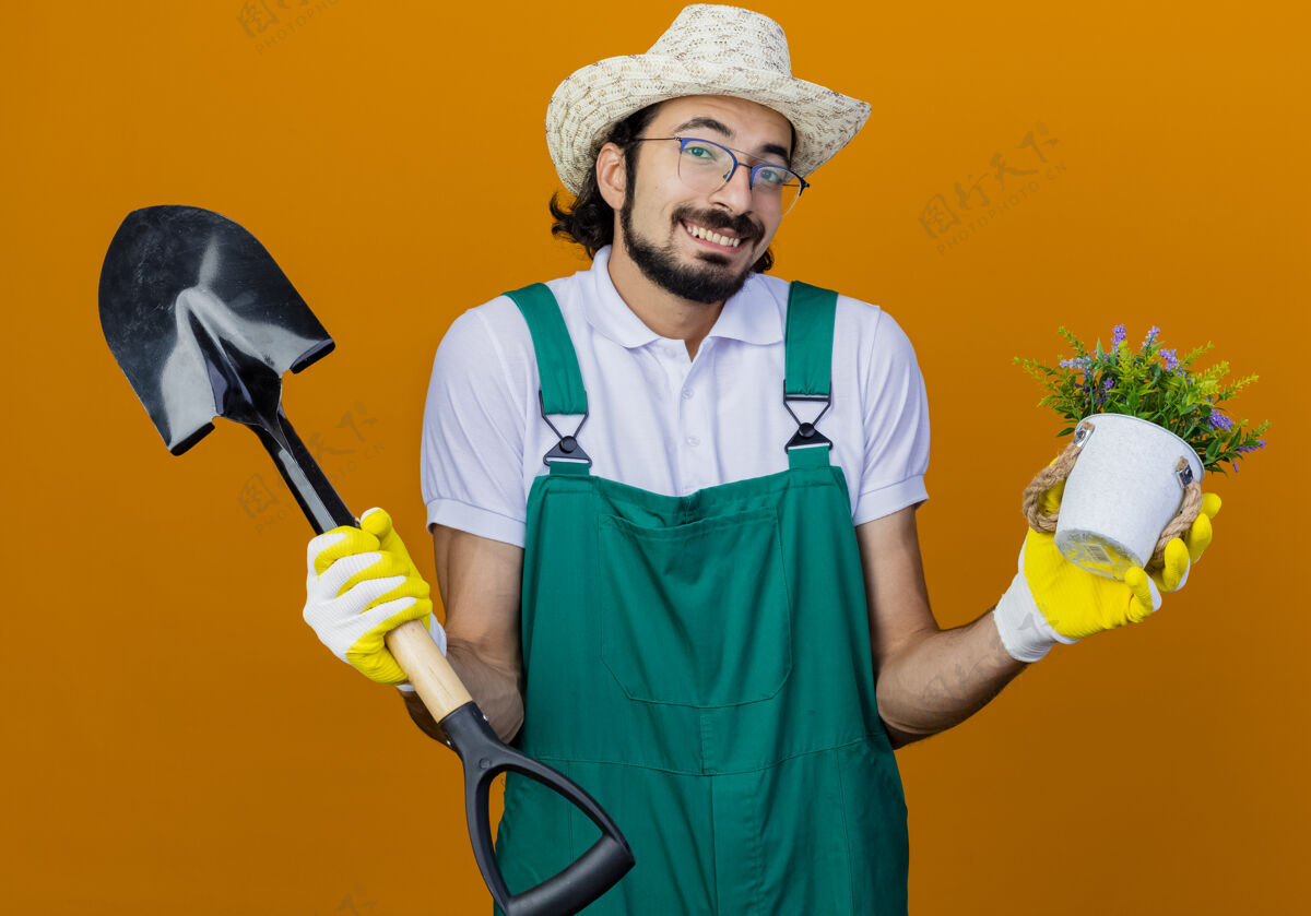 男人年轻的留着胡须的园丁 穿着连体衣 戴着帽子 拿着铲子和盆栽植物 困惑地耸耸肩穿着耸肩植物
