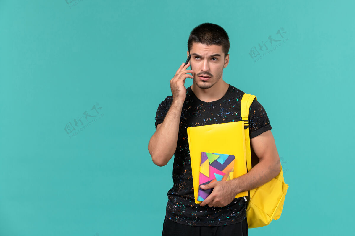 学院前视图中的男学生穿着深色t恤黄色背包拿着文案笔和文件在浅蓝色的墙上背包人持有