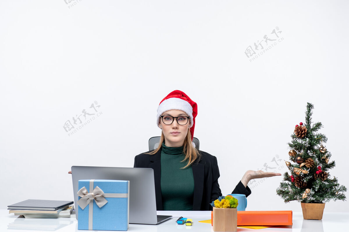 积极积极自信的女商人 戴着圣诞老人帽 坐在一张桌子旁 桌子上有圣诞树和一份白色背景的礼物笔记本电脑工作树