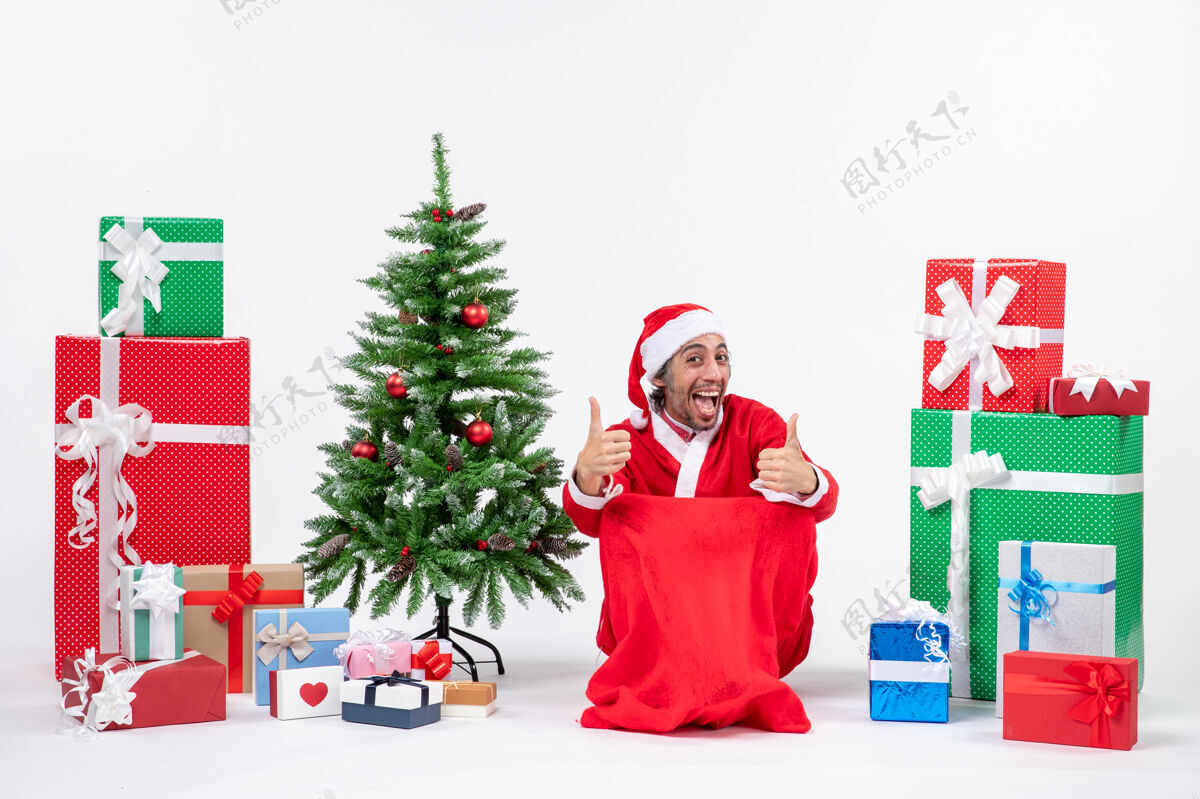 礼物情绪激动的年轻人打扮成圣诞老人与礼物和装饰圣诞树作出完美的姿态在白色背景情感圣诞老人礼物