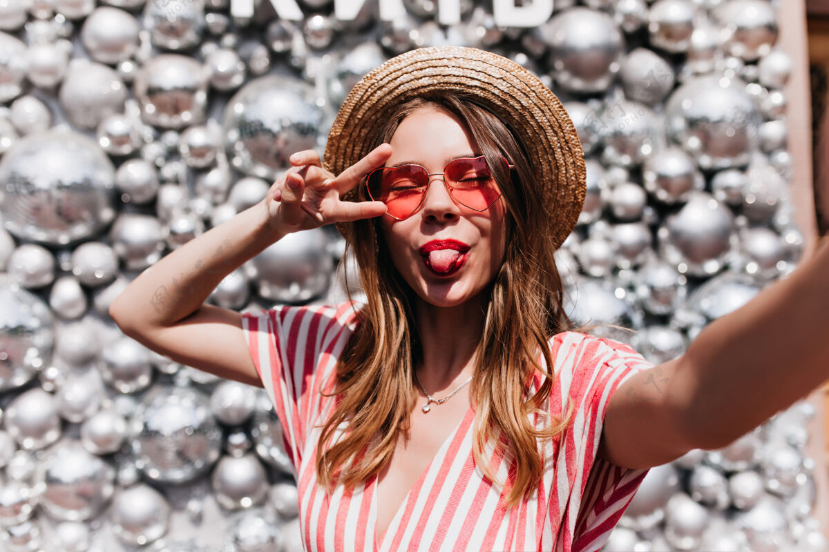配饰戴着粉色太阳镜的迷人年轻女子在自拍戴草帽的可爱女孩在迪斯科舞会附近摆出舌头的姿势表情女性人物