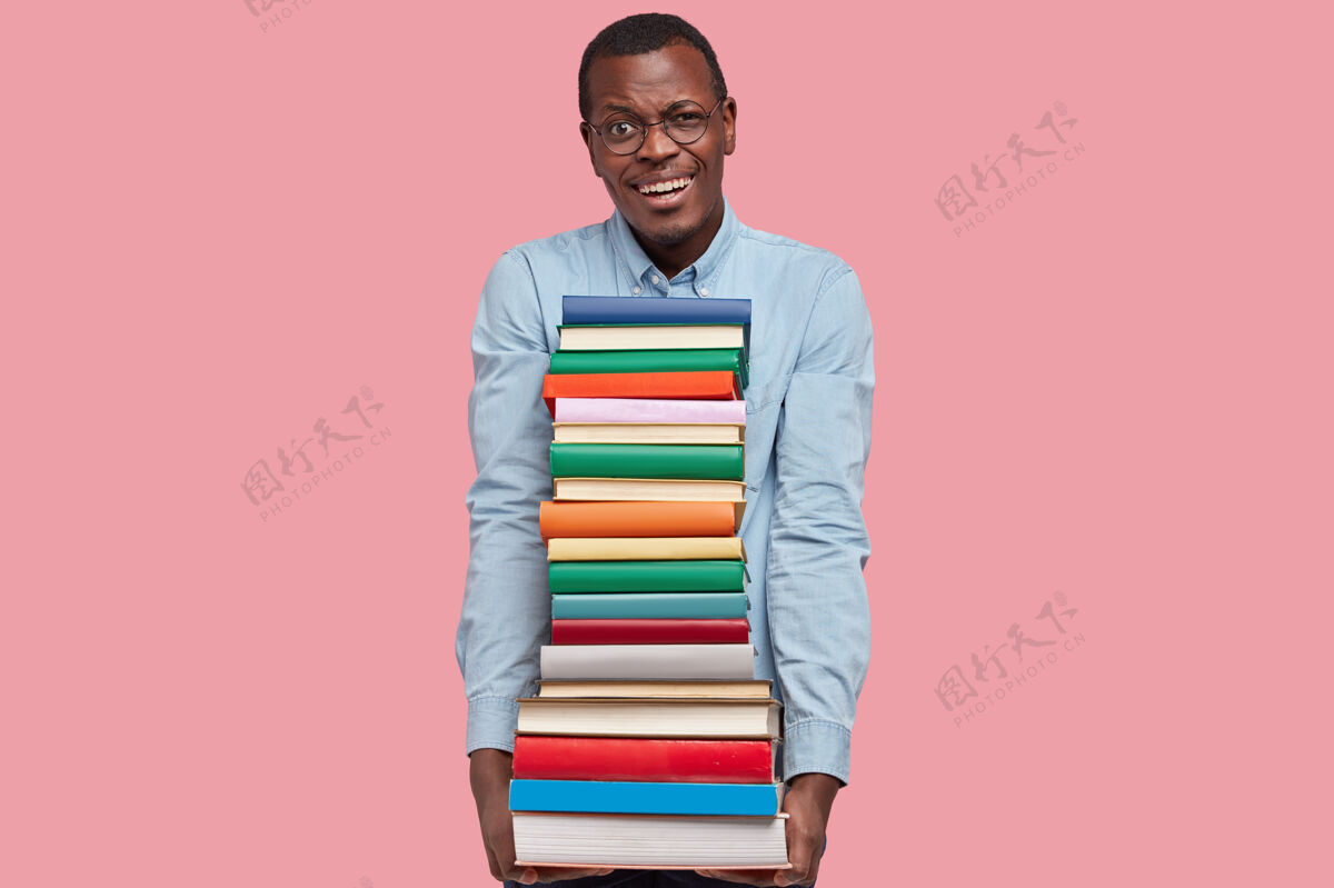 学生正面满意的黑皮肤男人 露出满意的表情 露出洁白的牙齿 双手拿着手册 穿着正式的衬衫堆眼镜书籍