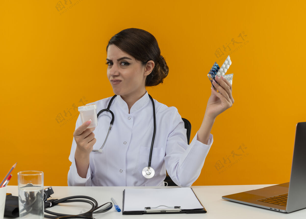 工具困惑的年轻女医生穿着医用长袍 手持听诊器 坐在办公桌旁 拿着医疗工具 拿着空罐子和隔离黄墙上的药片 在电脑上工作医生困惑穿着