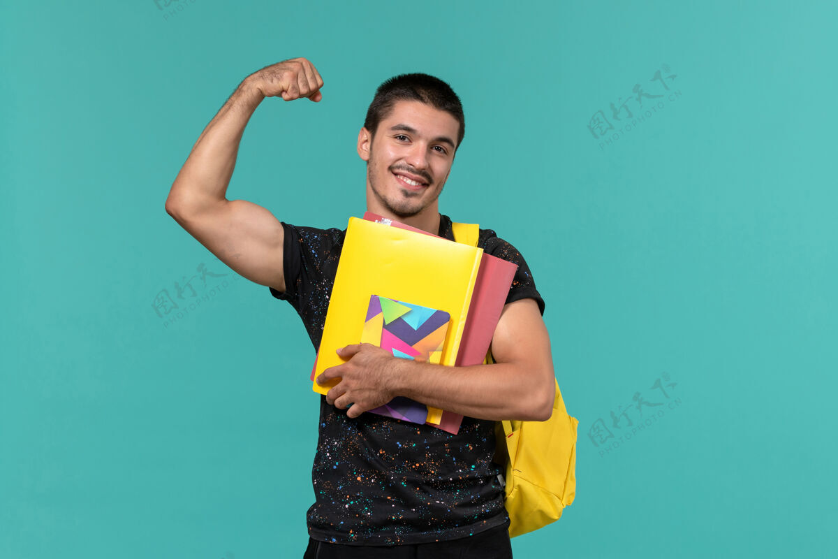 教训前视图中的男学生穿着深色t恤黄色背包 在浅蓝色的墙上拿着文件和抄写本大学快乐人