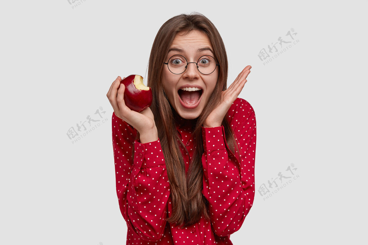 人类兴奋过度的快乐女士举手靠近脸 张大嘴巴 吃美味的苹果 戴透明眼镜饮食衬衫水果