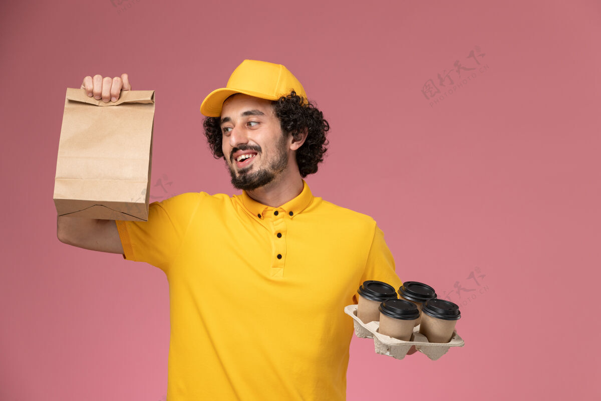 黄色正面图：身穿黄色制服的男性快递员手持棕色的咖啡杯和食品包装放在粉红色的墙上正面快递员微笑