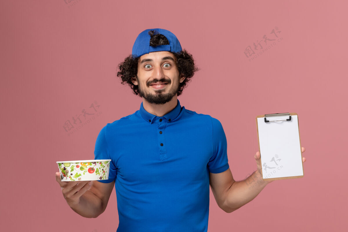 男性正面图：身穿蓝色制服 披风的男性信使拿着记事本和粉红色墙上的圆形投递碗碗成人正面